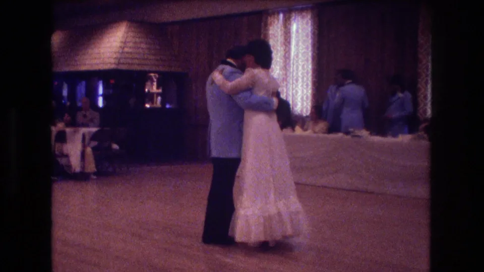 A closer home-movie view shows the bride and groom holding each other during their reception dance, with wood paneling, a bar or service area, and seated guests in the background.