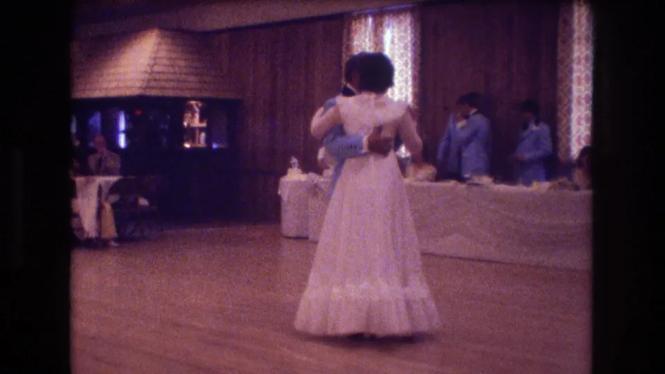 From behind the bride, the 1974 Boston reception footage shows the couple slow dancing while guests sit and stand around long tables under warm interior light.