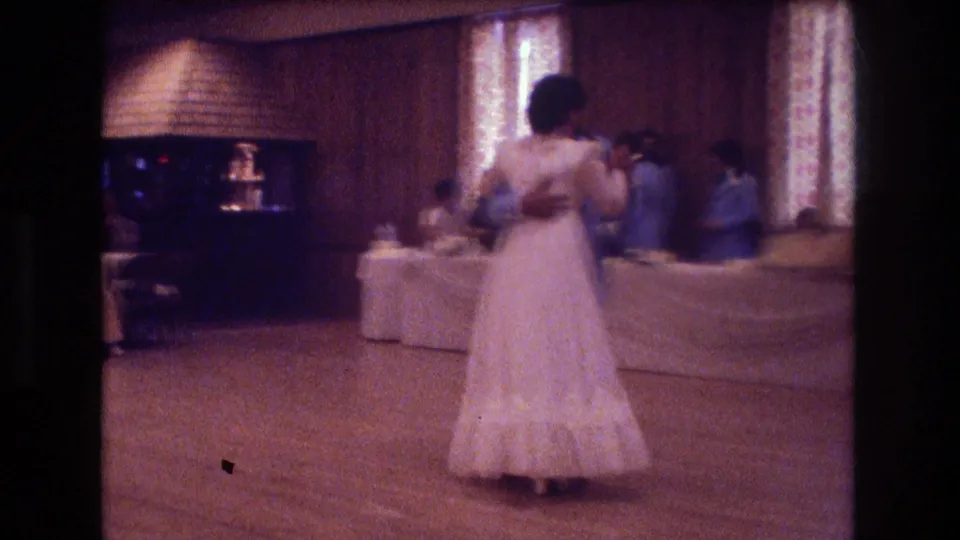 At a 1974 Boston wedding reception, a bride in a long white dress dances with a partner in a wood-paneled hall with patterned curtains and white-covered tables.