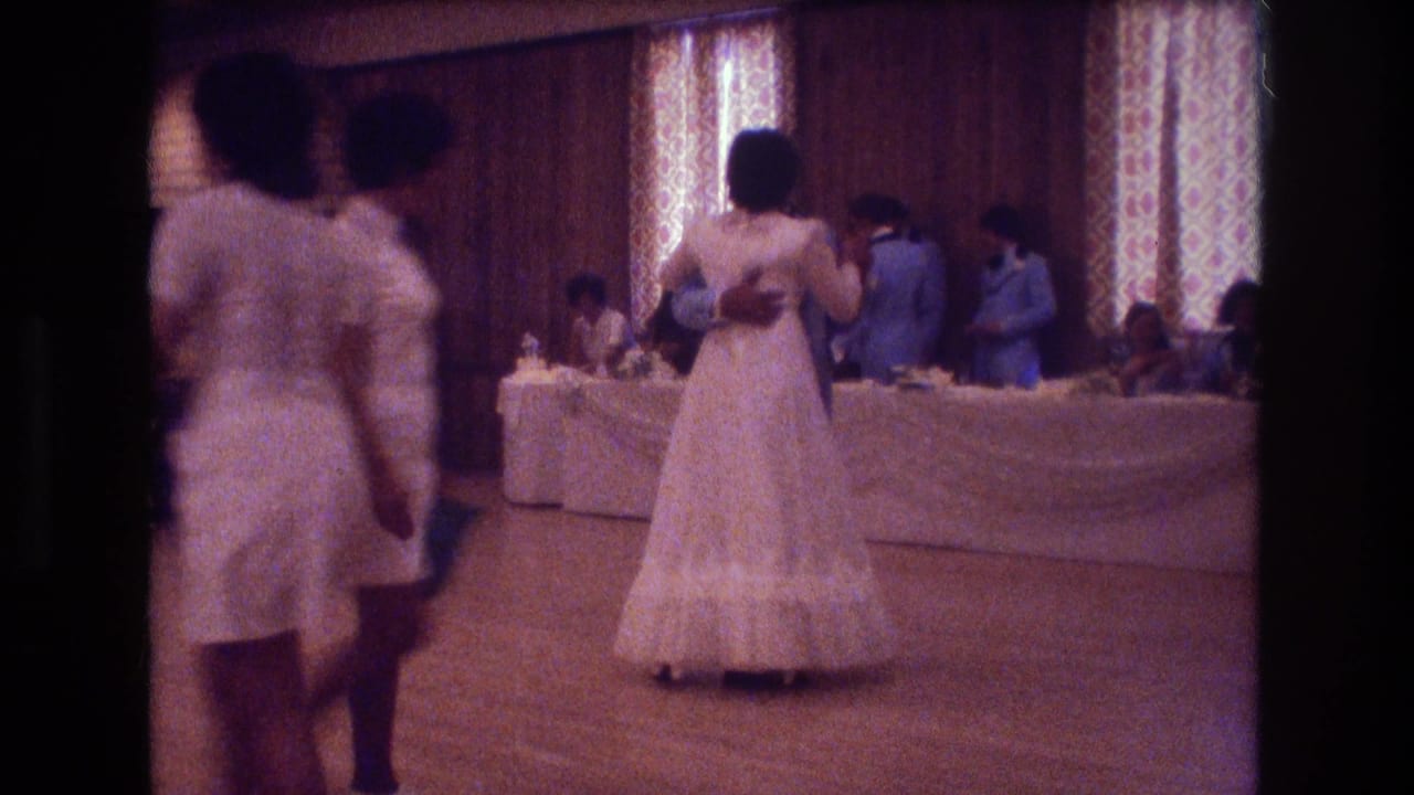 Guests stand near the dance floor as a bride dances in front of reception tables and patterned curtains.