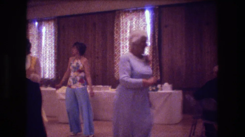 At the wedding reception, a woman in a pale dress and another in a floral top with wide-leg pants move across the dance floor in a 1970s interior.