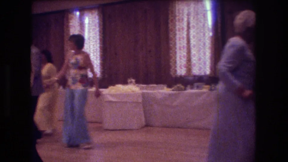 Several guests at the 1974 Boston wedding reception stand or walk near the dance floor, including a woman in a floral top and wide-leg pants and another in a pale dress.