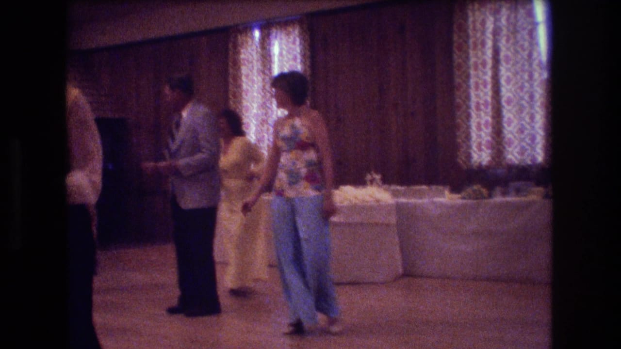 In this supported 1974 Boston home-movie frame, adults dance in colorful 1970s clothing near buffet tables in a warmly lit wood-paneled hall.