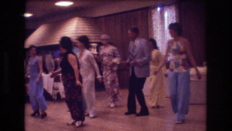Adults dance in a Boston indoor hall, with flared pants, patterned dresses, seated guests, and a large hooded wall feature visible in the background.