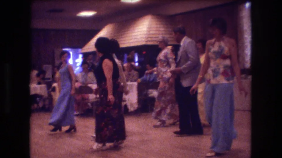 A group of adults dances in a warm indoor hall, with seated guests, tables, wood paneling, and colorful 1970s outfits visible around them.