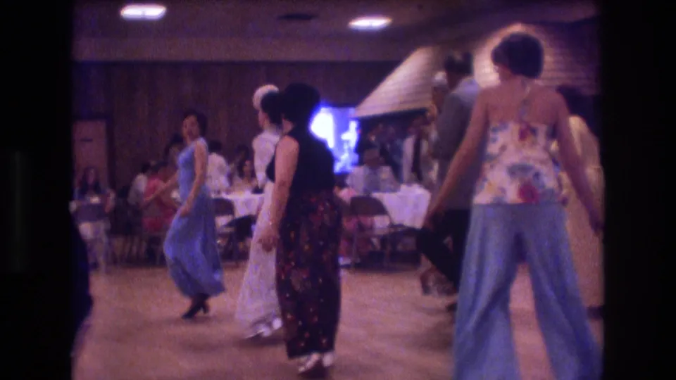 Adults continue dancing across the hall floor in the 1974 Boston home-movie footage, with seated guests, buffet or dining tables, wood paneling, and bright window light behind them.