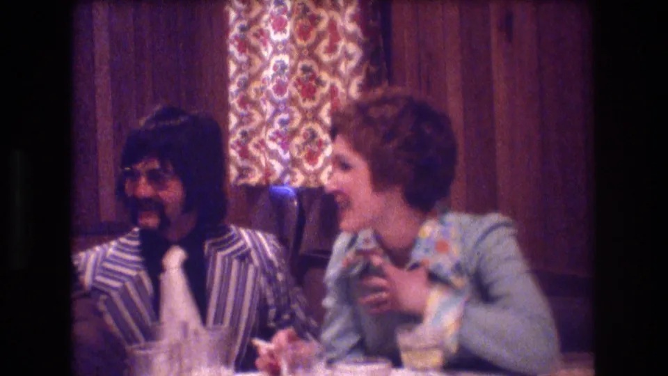 A mustached man in a striped suit and a woman in a light blouse sit at a table in a private interior, talking during the 1974 Boston gathering described by the metadata.