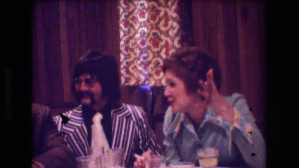 The seated pair continue an animated conversation at the table, with drinks and patterned wall decor visible in the 8mm home-movie frame.