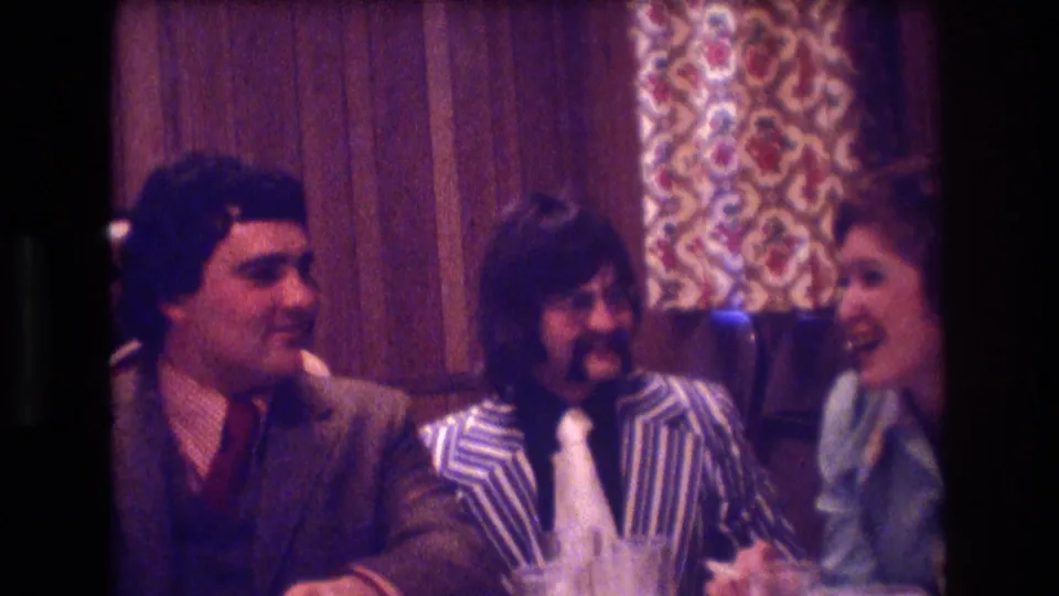 Three adults in 1970s formal attire sit at a warmly lit Boston restaurant table, including a man in a striped jacket and a woman leaning in conversation.