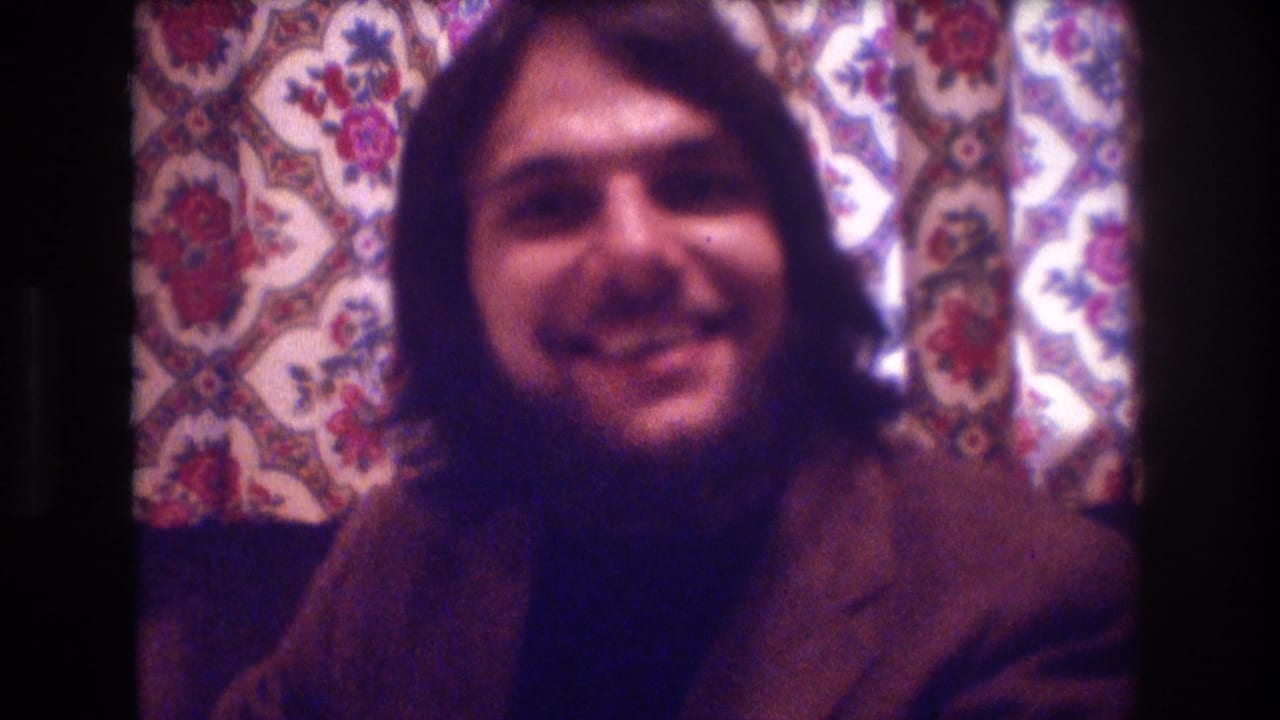 Smiling man in a brown jacket seated in front of floral curtains.
