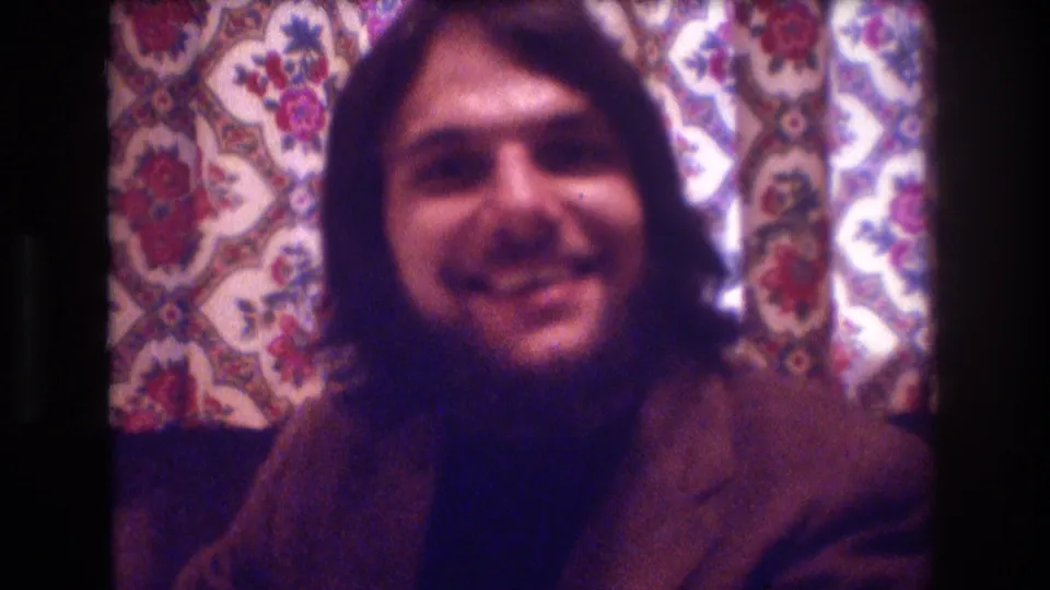 A smiling long-haired man in a brown jacket faces the home-movie camera inside the 1974 Boston restaurant interior.