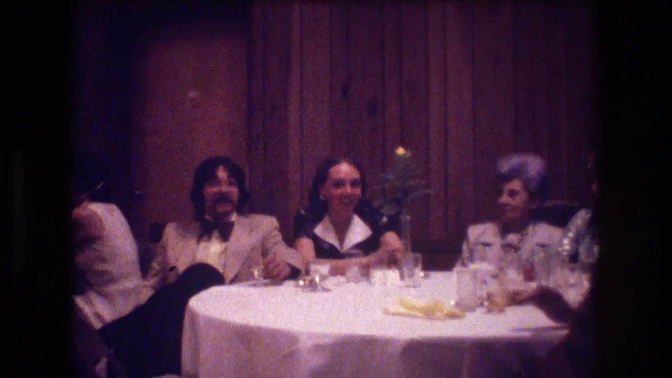 Several adults sit around a round white-clothed restaurant table in Boston, with 1970s clothing, glassware, plates, and wood-paneled walls visible.