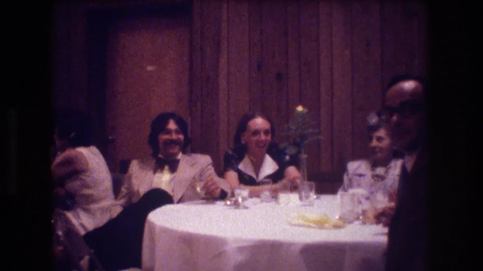 Adults continue dining and talking at a 1974 Boston restaurant table, with a man in a light jacket, women seated nearby, and a man with glasses at the right edge.