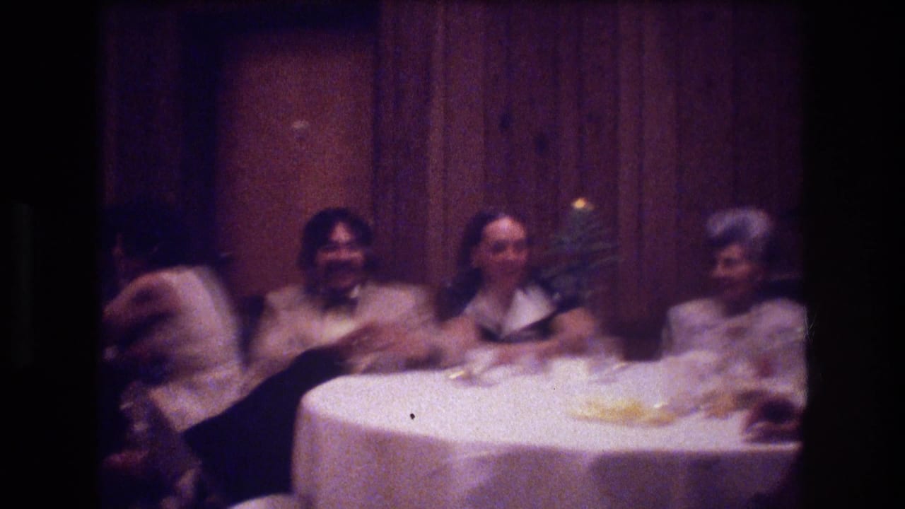 Boston 1974 home-movie frame showing formally dressed wedding guests seated around a white-clothed dinner table in a wood-paneled reception interior.