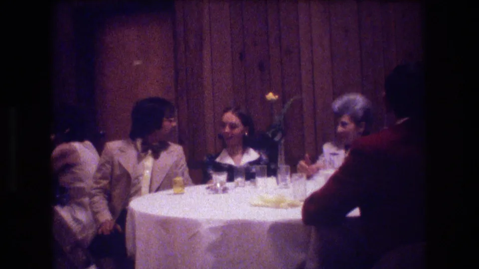 Boston 1974 frame of wedding guests in formal attire conversing at a round table with glasses and a small floral centerpiece visible.