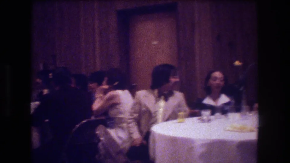 Boston 1974 home-movie frame showing seated reception guests in profile and partial view as they talk beside a white tablecloth and wood-paneled wall.