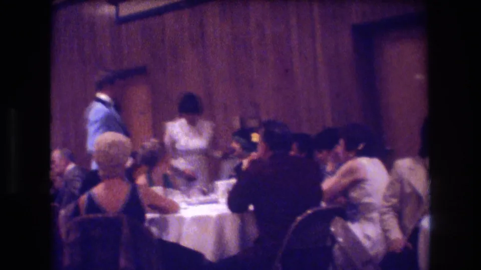 Boston 1974 frame showing a wider wedding reception room with seated guests, a server in a light uniform, white tablecloths, and wood-paneled walls.