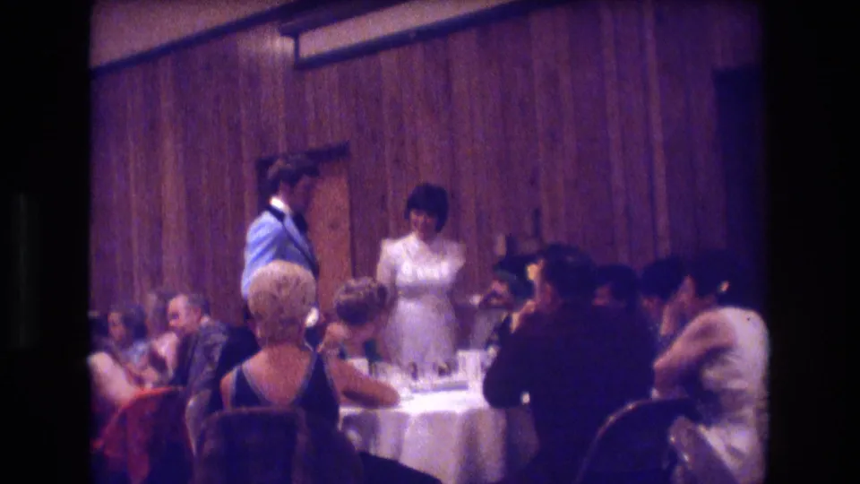 In the supplied 1974 Boston wedding reception footage, the bride in a white gown and groom in a light blue tuxedo stand near guests seated at a round table in a wood-paneled banquet room.