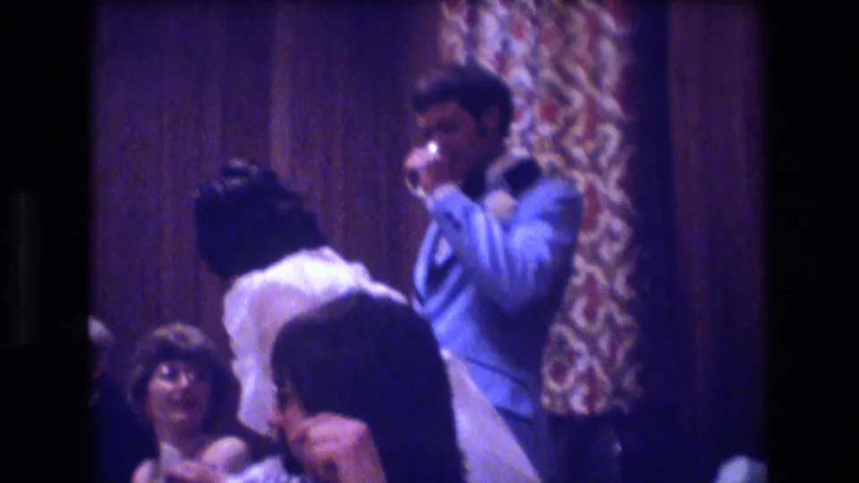 The groom appears to drink or toast while standing beside seated guests in the wood-paneled reception room shown in the 1974 Boston home-movie clip.