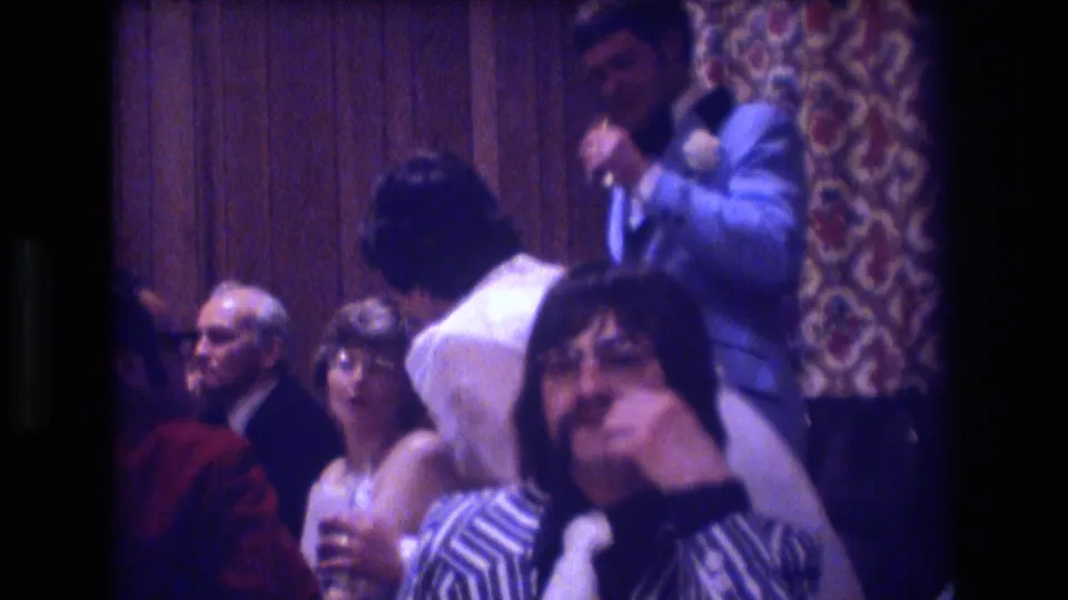 Several guests face the camera while the groom and bride stand nearby in the banquet-hall interior of the supplied 1974 Boston wedding reception footage.
