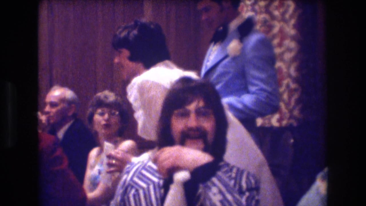 1974 Boston 8mm frame showing seated adults at an indoor party, including a man in a striped shirt with glasses and mustache facing the camera while other guests sit and stand nearby.