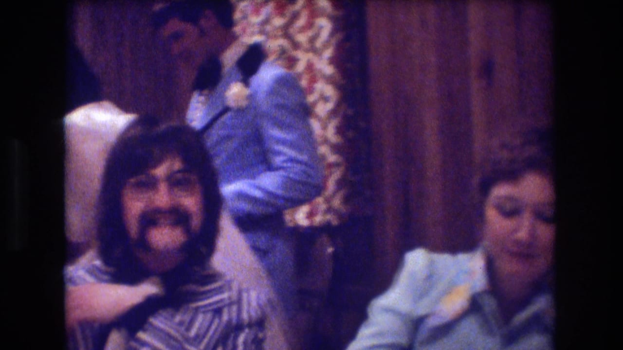 1974 Boston home-movie frame with a man in glasses and mustache smiling at the camera while a woman in a pale blue blouse sits nearby and a suited guest stands in the background.