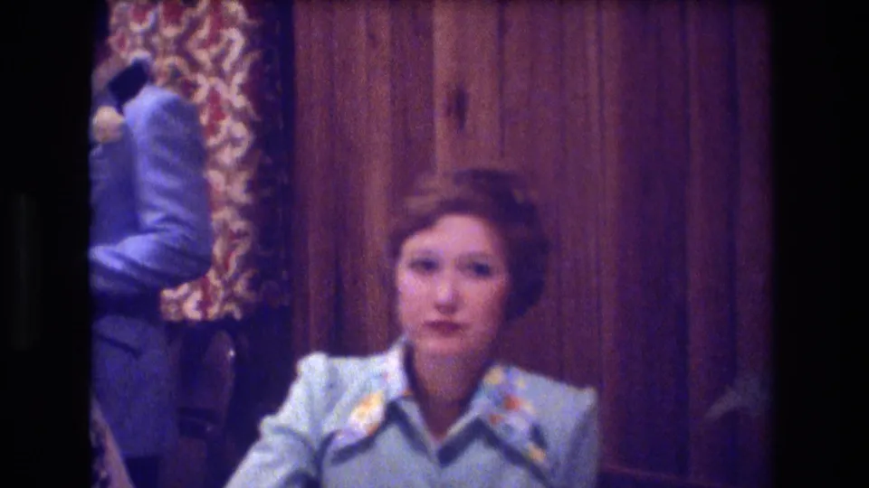 1974 Boston 8mm frame showing a woman in a pale blue patterned blouse seated in a wood-paneled party room, with part of a man in a blue suit visible at the left edge.