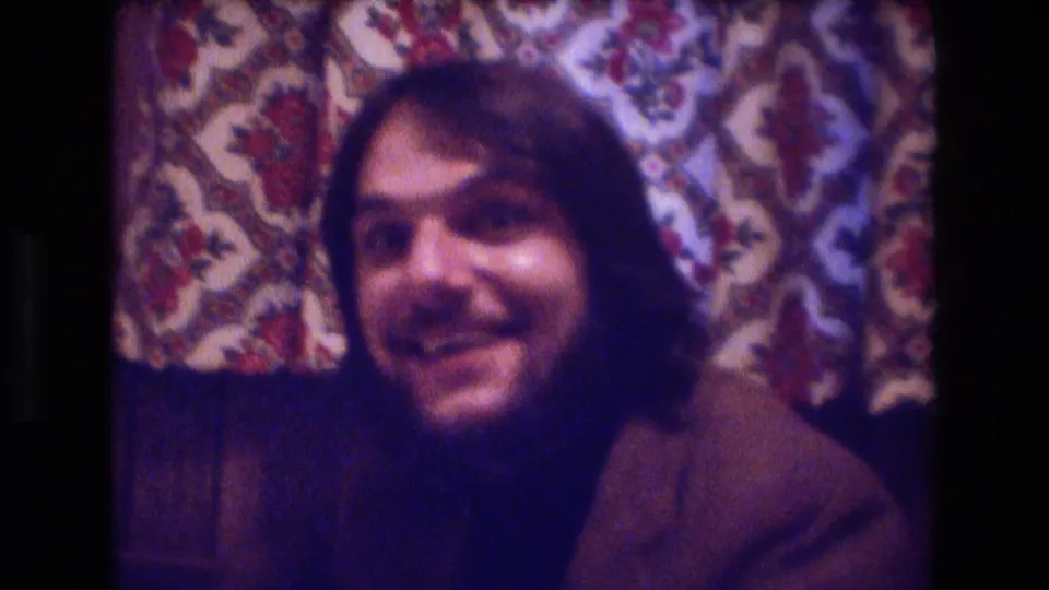 1974 Boston home-movie close-up of a long-haired man with facial hair smiling toward the camera in an indoor party setting with patterned wallpaper behind him.