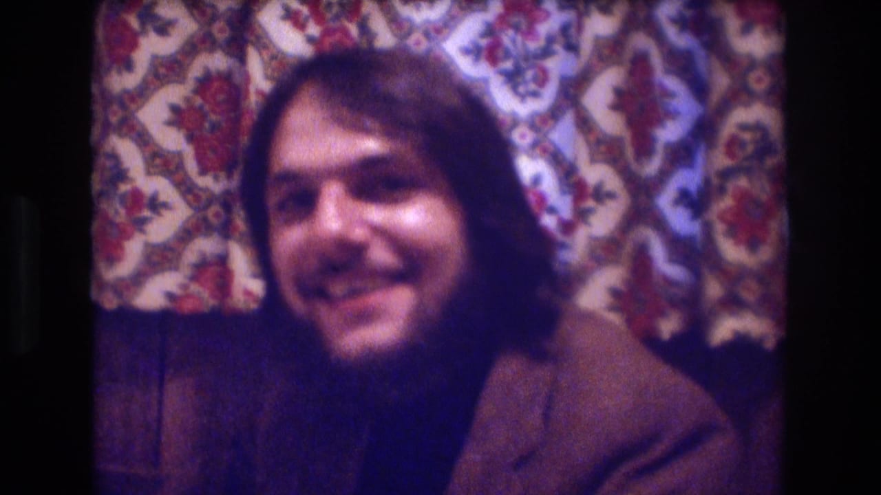 In a 1974 Boston home-movie frame, a long-haired bearded man smiles toward the camera beside a patterned curtain in a wood-walled room.