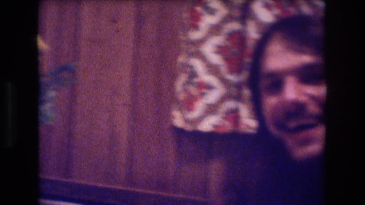 Partial view of a smiling bearded man beside wood paneling and patterned fabric.