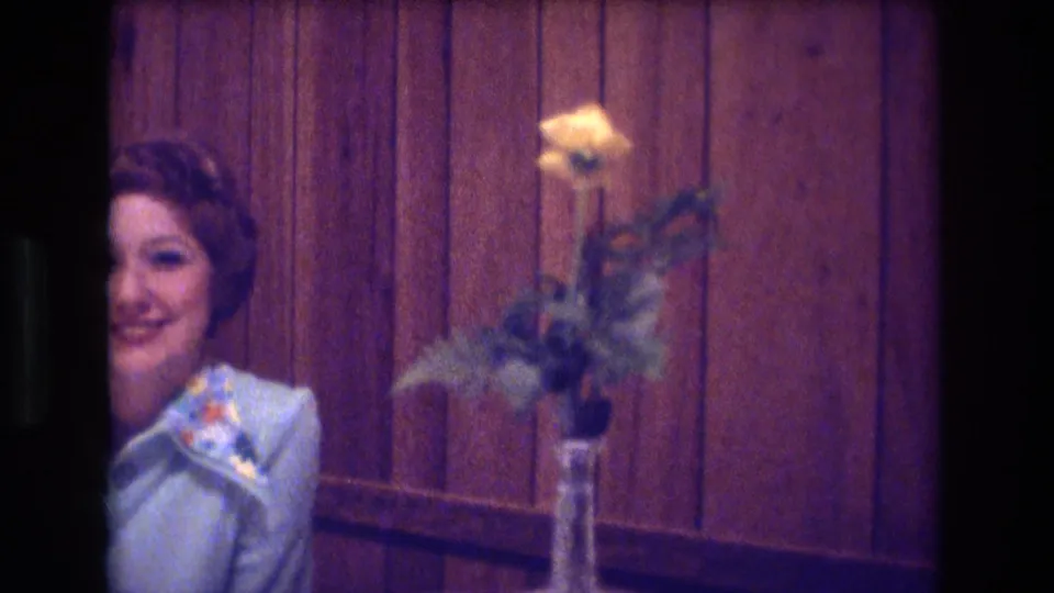 A woman in a light blue blouse is partly framed near a small table holding a vase with a yellow rose against wood-paneled walls.