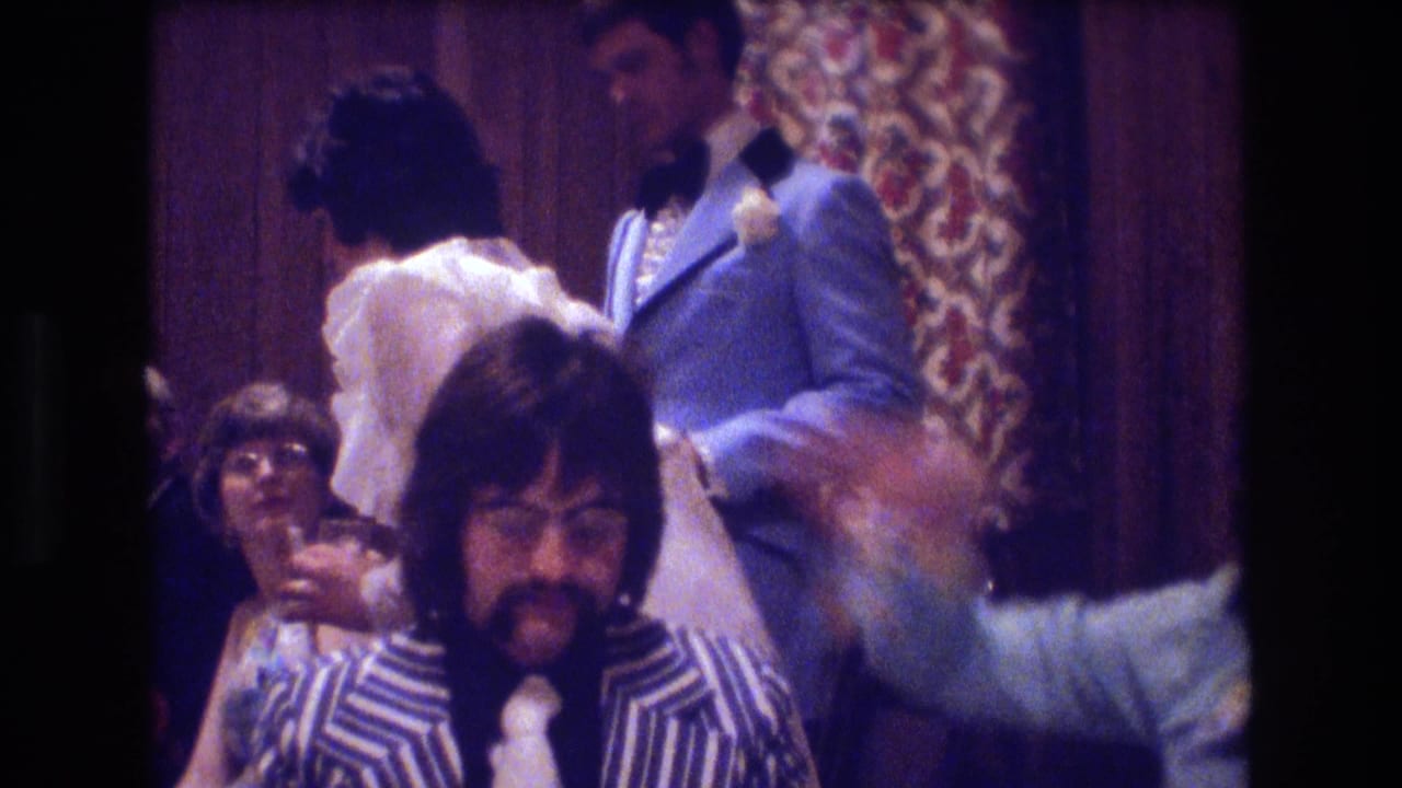 Man in a striped jacket sits near dancing guests and a man in a light blue tuxedo.