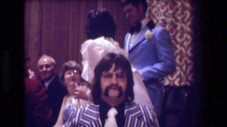 A seated man in a blue and white striped jacket looks toward the camera while formally dressed party guests stand and move behind him in the 1974 Boston footage.
