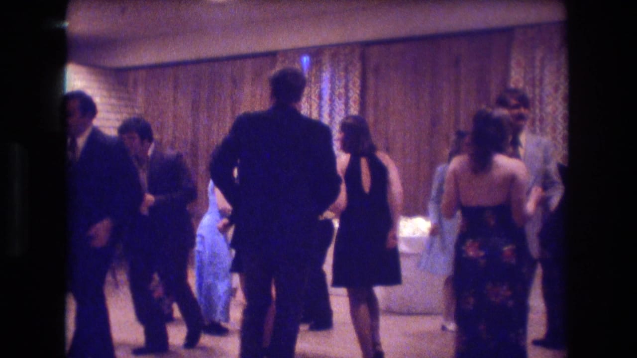 Adults in suits and dresses dance in a dim indoor party room.