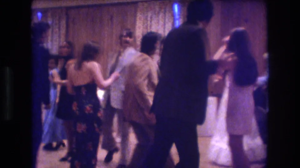 A crowded 1974 Boston home-movie party frame shows adults in formal and patterned clothing moving across a dim dance floor.