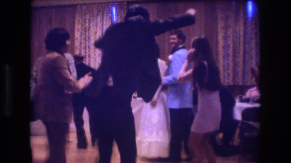 Adults at a 1974 Boston indoor party dance and gesture near the edge of the room, with tables and seated guests partly visible in soft 8mm footage.