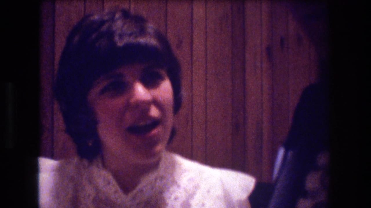 A young woman in a white formal dress appears to speak during a 1974 Boston indoor social gathering, with wood-paneled walls behind her.