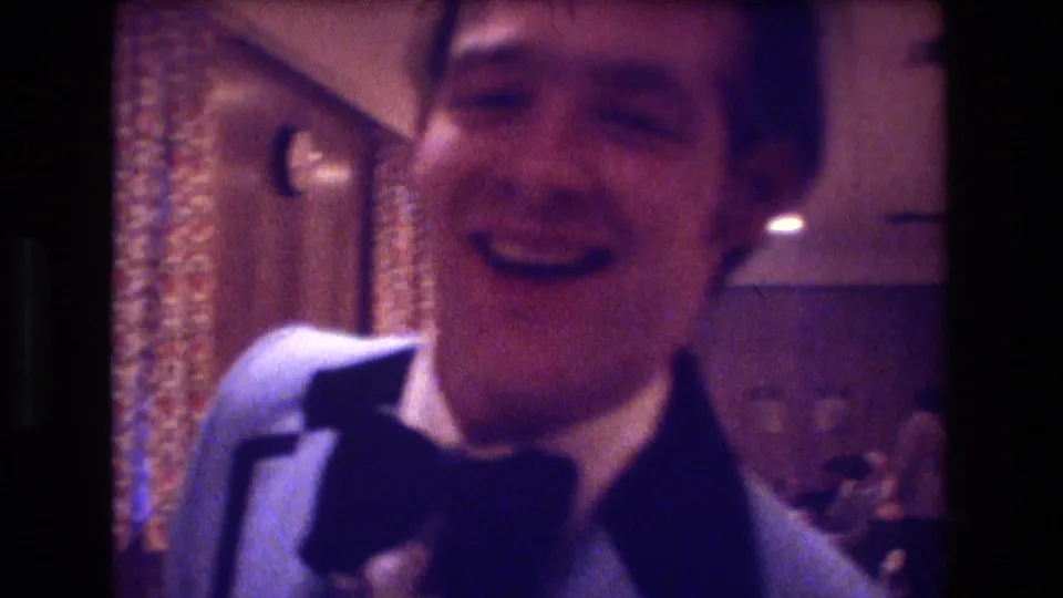 A young man in a light blue tuxedo jacket laughs in a close, grainy 8mm frame from the 1974 Boston formal gathering.
