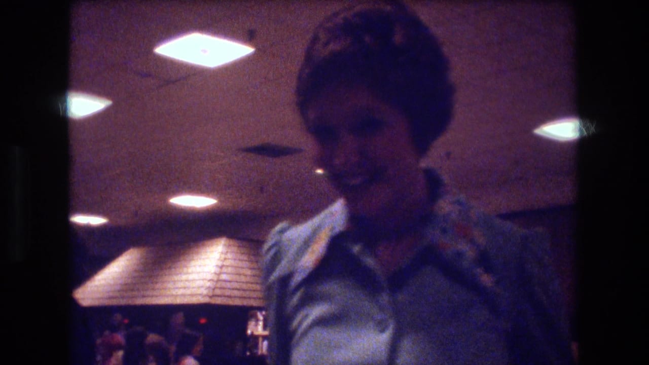 In 1974 Boston home-movie footage, a woman in a light blue patterned blouse stands near the camera under ceiling lights inside a reception hall.