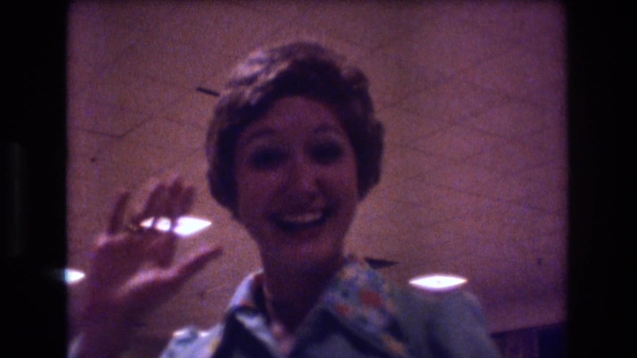 A smiling woman in a light blue patterned blouse appears to wave toward the camera during a 1974 Boston indoor wedding reception.