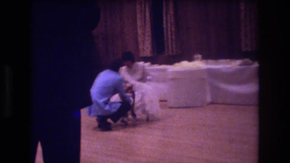 A man crouches beside a bride in a white wedding dress near a long white-draped table in an indoor Boston reception setting from 1974.