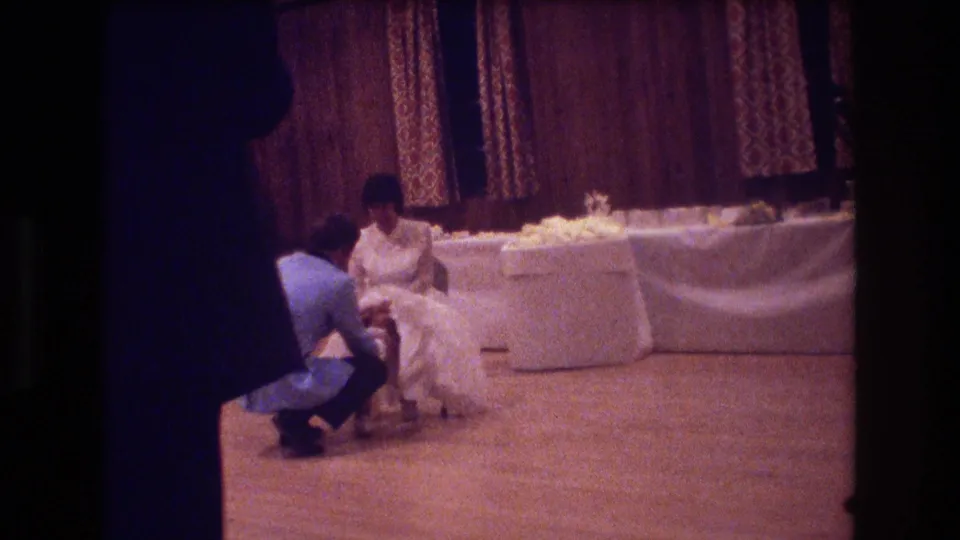 The groom appears to adjust the bride's garter as she sits near a white-draped table in a warm-toned 1974 Boston wedding reception scene.