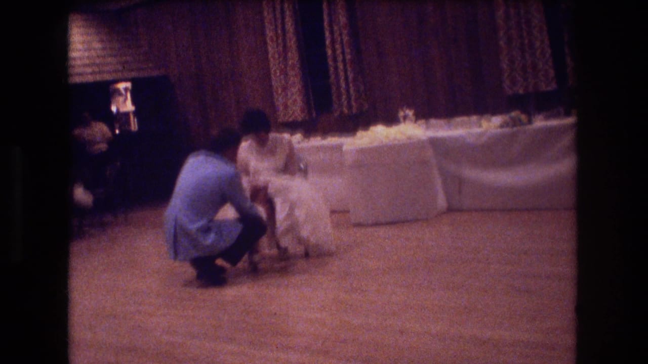 In a 1974 Boston wedding reception scene, a groom in a light blue suit kneels before a bride in a white dress near a long linen-covered table.