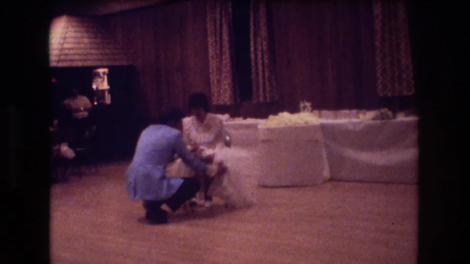 The groom remains crouched in front of the bride during the garter-removal tradition, with reception linens and curtains visible behind them.