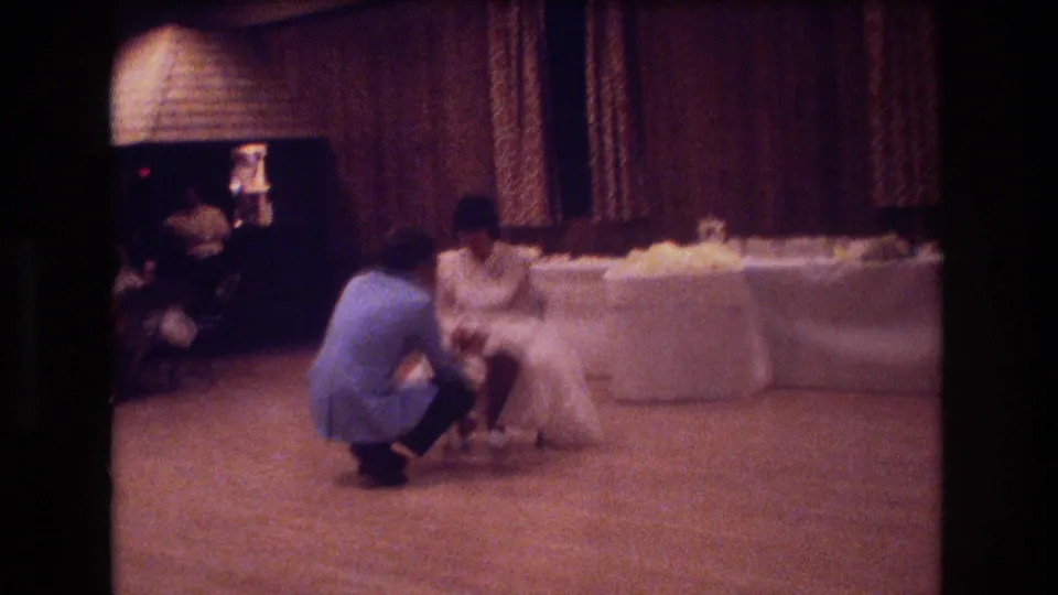 The bride sits near the head table while the groom kneels low in front of her, framed by warm interior lighting and 1970s reception decor.