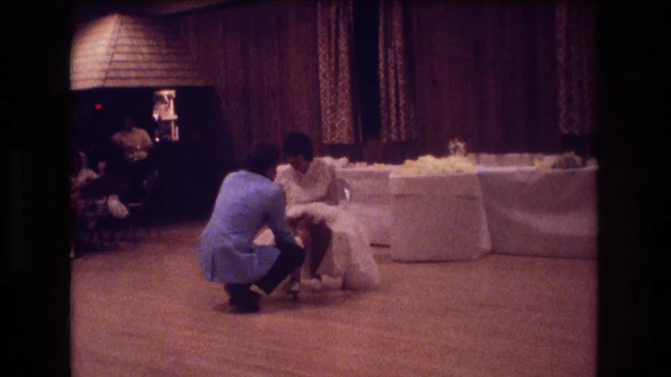 The 8mm frame shows the groom still kneeling before the bride at the Boston wedding reception, with the long table and patterned curtains behind them.
