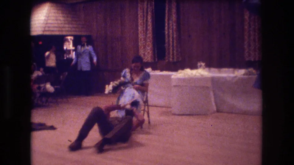 A party guest sits on the floor near draped banquet tables as other guests stand by wood-paneled walls in this 1974 Boston home-movie frame.
