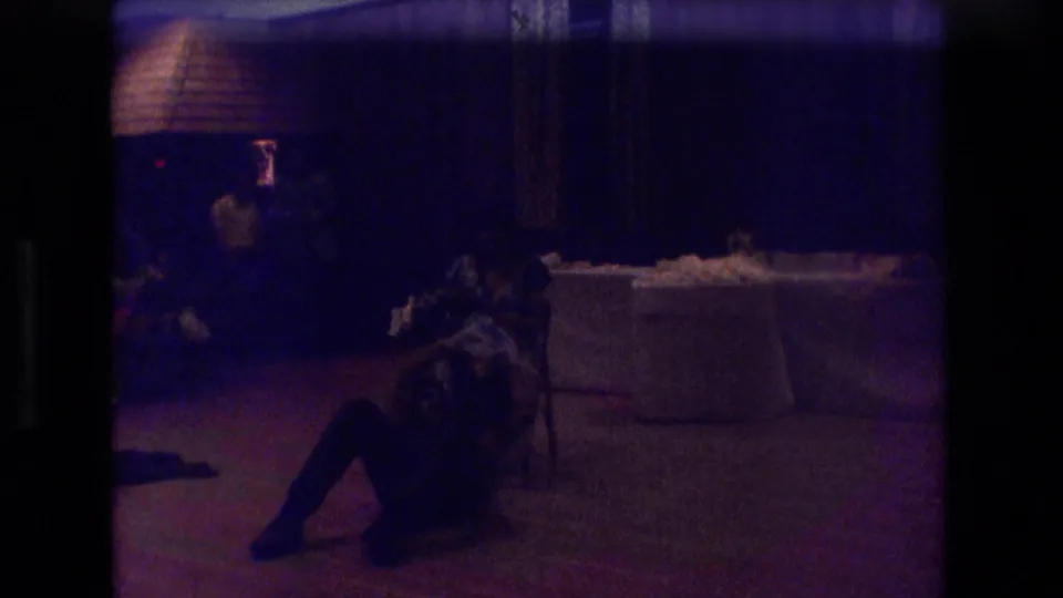 A darker 8mm frame shows a seated party guest in shadow near draped tables and curtains inside the 1974 Boston indoor gathering.