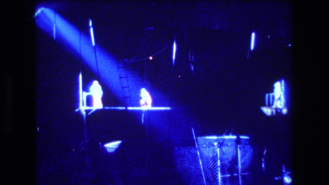 1974 Boston home-movie frame showing a dark circus performance area under blue stage lighting, with a spotlight, platform, fencing, and distant figures near the ring.