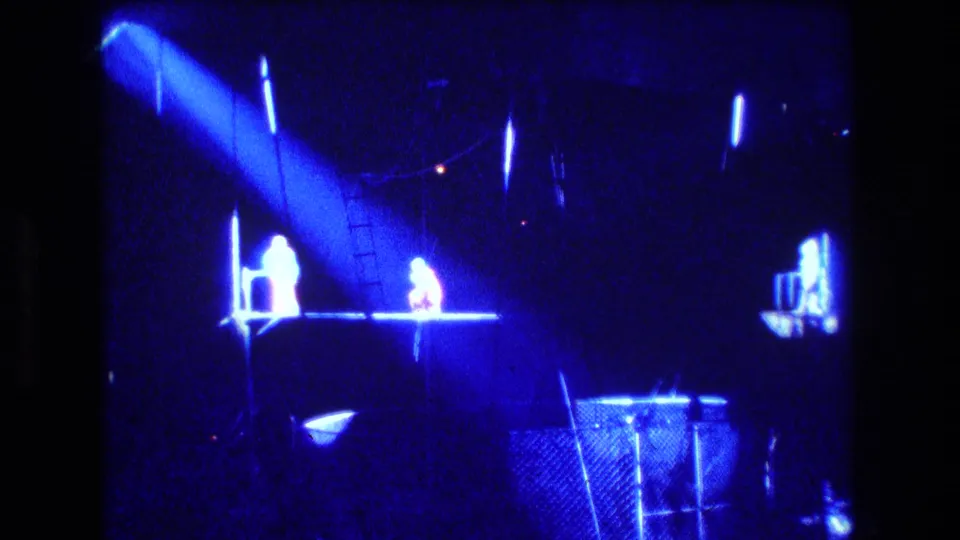 1974 Boston home-movie frame showing a dark circus performance area under blue stage lighting, with a spotlight, platform, fencing, and distant figures near the ring.